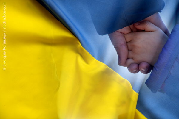 An adult holding the hand of a child with the Ukrainian flag on the background