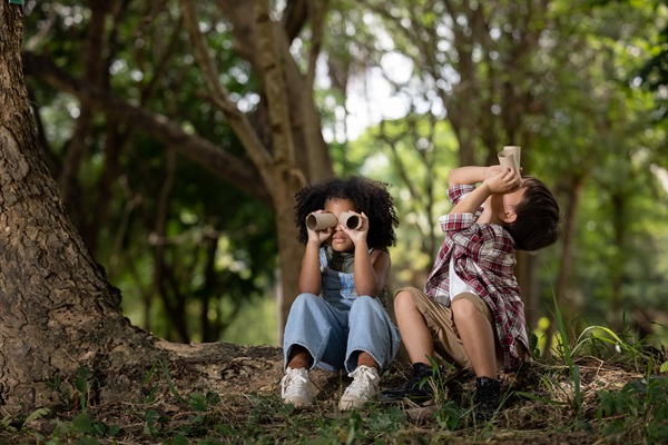 Two children sitting in forest and using toilet paper rolls as binoculars