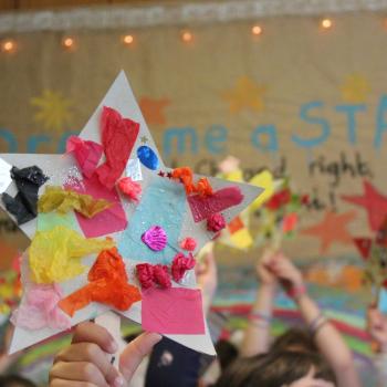 Image of students in front of a large rainbow artwork.  The students are holding decorated star wands above their heads.
