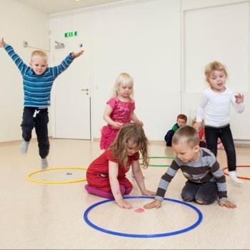 Photo of kindergarten children playing a core PTLM game. Two children are visiting a hoop with a letter in it. Other children are travelling to the hoop. The are all engaged in the game.