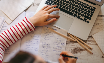 Student writing on a paper notebook while using a laptop