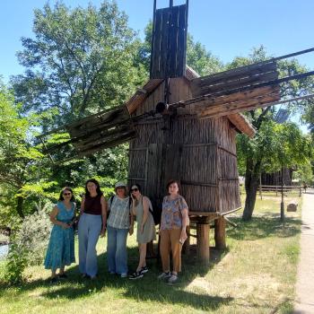 A group of teachers stands in front of a traditional wooden windmill at the Village Museum in Bucharest during an outdoor educational activity.