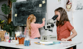 two students in a classroom: One using a microscope, the other wriiting on the blackboard