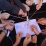 many student's hands each hodling a thread that is ties to one marker in the centre and trying to write with it