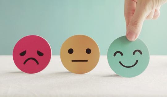 Smiley, neutral, and frowny faces made of paper, smiley being chosen by a hand