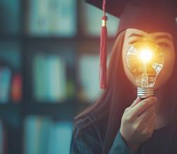 Woman in graduation gown and hat holding lit lightbulb