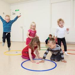 Photo of kindergarten children playing a core PTLM game. Two children are visiting a hoop with a letter in it. Other children are travelling to the hoop. The are all engaged in the game.