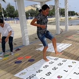 Teachers enjoying active learning on a wooden platform on the beach. One teacher in the foreground is jumping and one in the background is reading instructions.