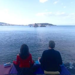 Two people meditating in front of the sea. In the background there is the town of Mytilene, in Lesvos island.