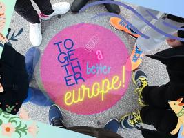 "A group of children is presented, only their feet are visible, standing in a circle. In the center of the circle, there is the inscription 'Together for a better Europe.'"